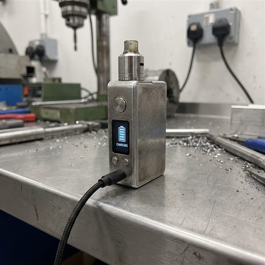 A chunky box-mod style AIO vape device connected to USB-C, OLED display reading CHARGING